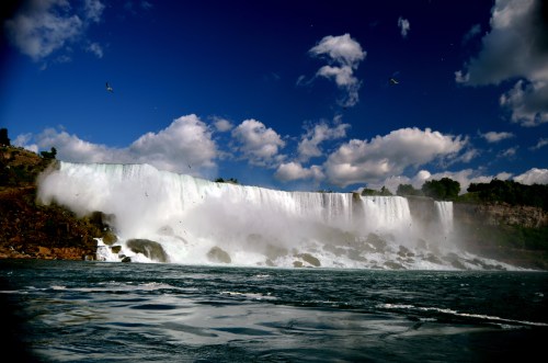 A majestic waterfall cascades over rocky cliffs, surrounded by mist and a blue sky with scattered clouds.