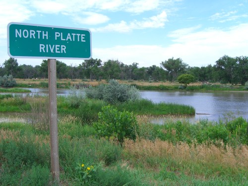 Sign for North Platte River beside a calm waterway surrounded by greenery and tall grasses under a blue sky.