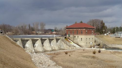 A concrete dam with a red-roofed building beside it, set against a cloudy sky and bare landscape.