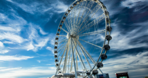 A large Ferris wheel against a blue sky with clouds, showcasing its structure and surrounding area.