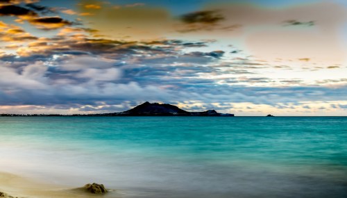 A serene seascape featuring calm turquoise waters, a distant island, and a colorful sky with clouds at sunset.