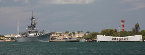 A battleship docked near a memorial structure, with palm trees and a lighthouse in the background.