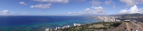 Panoramic view of a coastal city with blue ocean, green land, and scattered clouds under a bright sky.