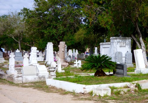 A peaceful cemetery scene with various gravestones, greenery, and trees under a partly cloudy sky.