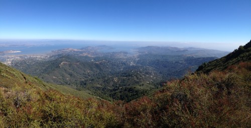 Panoramic view of rolling green hills and valleys, with a distant bay and mountains under a clear blue sky.