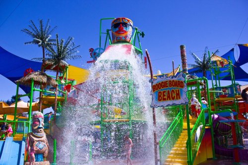 Colorful water park with a large water slide, splashing water, and tropical-themed decorations under a clear blue sky.