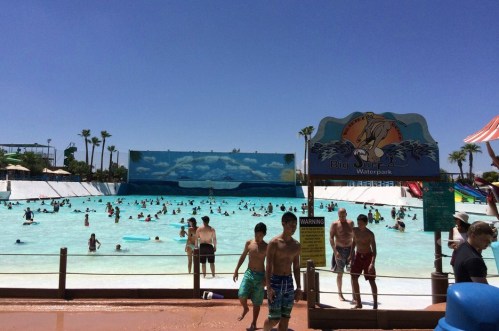 A busy waterpark with a wave pool, people swimming, and colorful slides under a clear blue sky.