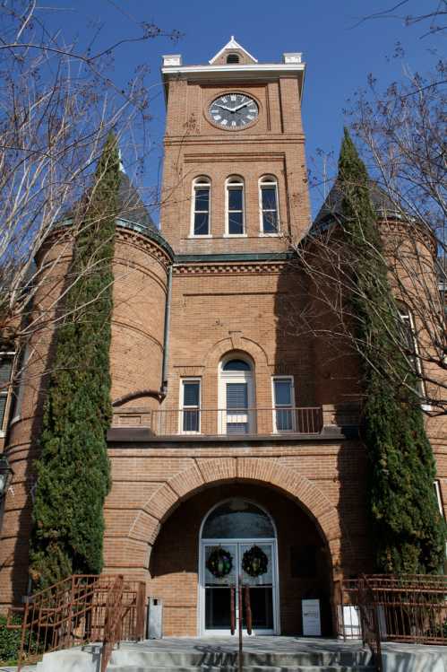 Historic brick building with a clock tower, flanked by tall trees, under a clear blue sky. Wreaths adorn the entrance.