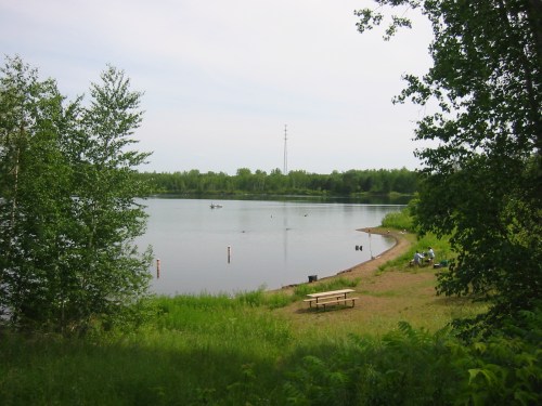 A serene lake surrounded by greenery, with a sandy shore and a picnic table, under a clear sky.