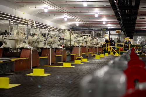 Industrial facility interior with machinery, yellow safety markings, and workers in hard hats.