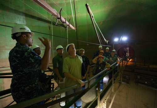 A group of people in hard hats observes and discusses inside a large, green industrial tunnel.