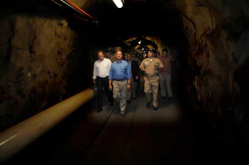 A group of men walking through a dimly lit tunnel, with pipes along the walls and a rugged, rocky interior.