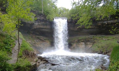 A cascading waterfall surrounded by lush greenery and rocky terrain, with mist rising from the water below.