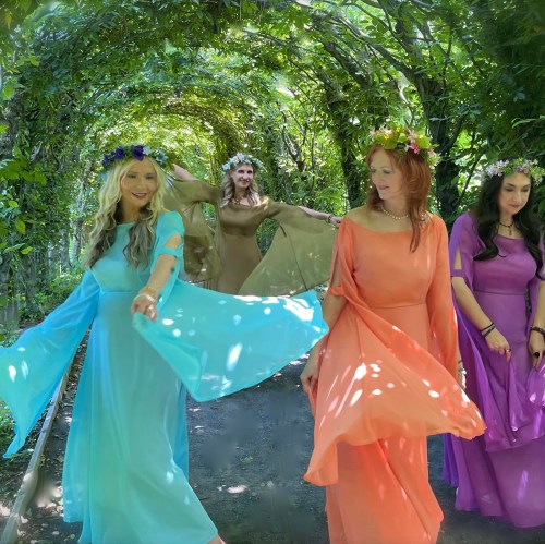 Four women in flowing, colorful dresses walk through a lush, green archway, adorned with flower crowns.