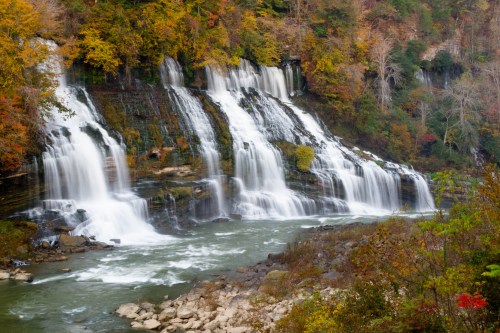 A serene waterfall cascades over rocky cliffs, surrounded by vibrant autumn foliage and a calm river below.