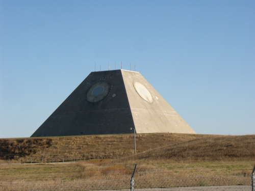 A large concrete pyramid structure with two circular openings, set against a clear blue sky and grassy landscape.