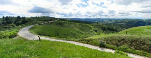 A winding road curves through lush green hills under a cloudy sky, showcasing a scenic landscape.