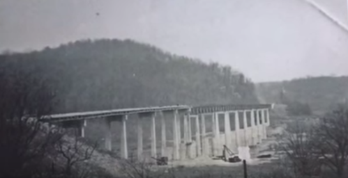 Black and white photo of a bridge under construction, supported by concrete pillars, with a hilly landscape in the background.