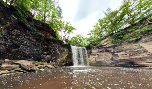 A serene waterfall cascades into a calm pool, surrounded by lush greenery and rocky cliffs.