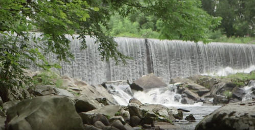 A serene waterfall cascades over a stone dam, surrounded by lush greenery and rocky terrain.
