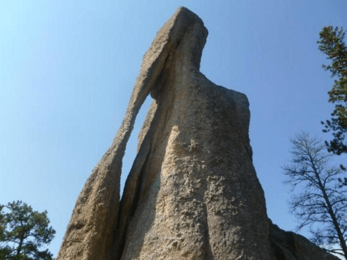 A tall, narrow rock formation with a distinct opening, set against a clear blue sky and surrounding trees.