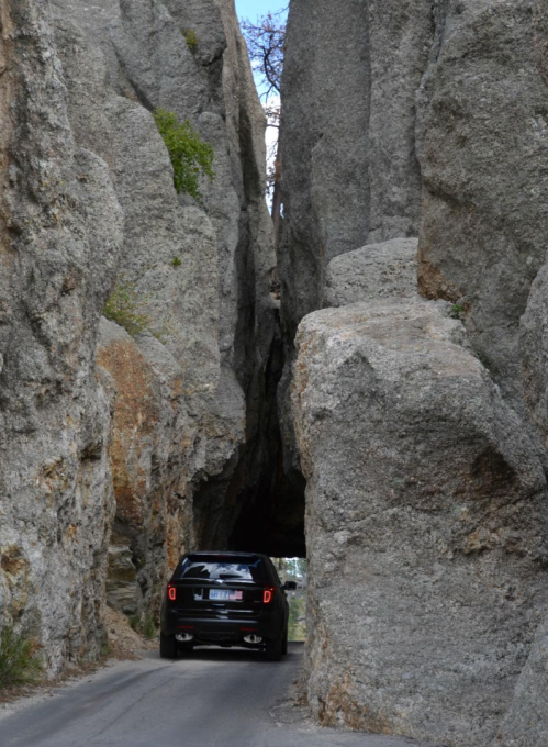 A black car drives through a narrow rocky passage between towering stone cliffs.
