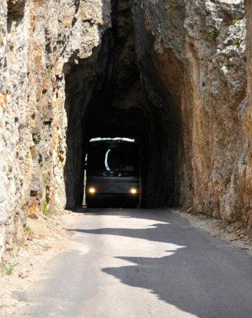 A bus drives through a narrow rock tunnel, surrounded by rugged stone walls and a clear blue sky above.