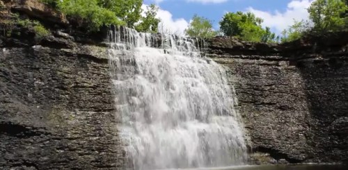 A cascading waterfall flows over rocky cliffs, surrounded by lush greenery and a bright blue sky.