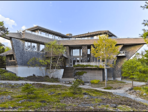 A modern, angular house with large windows, surrounded by greenery and a rocky landscape under a clear blue sky.