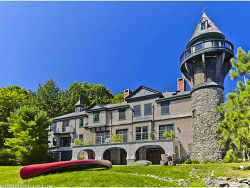 A large stone and wood mansion with a tower, surrounded by trees and a red canoe in front, under a clear blue sky.