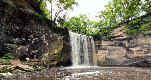 A serene waterfall cascades over rocky cliffs, surrounded by lush greenery and a calm pool below.