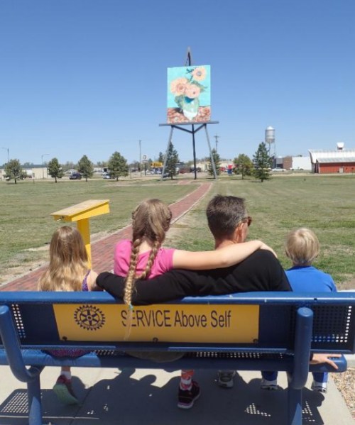 A family sits on a bench, looking at a large painting of flowers in a park on a sunny day.
