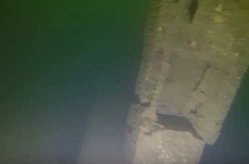 Underwater view of a submerged structure, partially covered in sediment, with greenish water surrounding it.