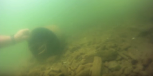 A submerged underwater scene showing a hand reaching towards a dark object on a rocky, sandy bottom.