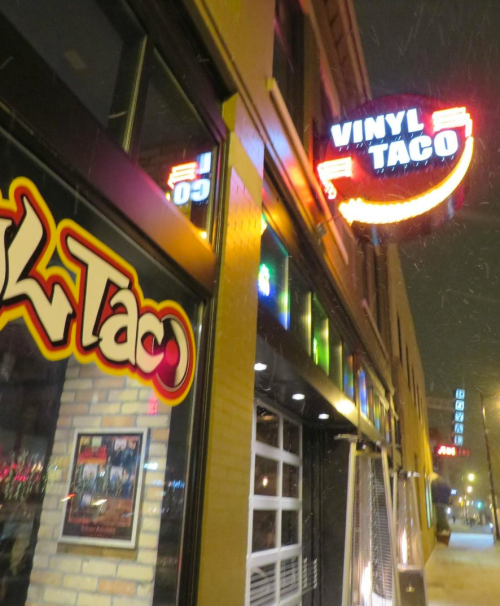 A brightly lit sign for "Vinyl Taco" outside a restaurant on a snowy evening.