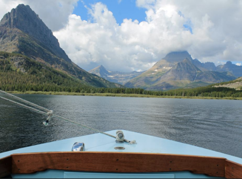 View from the bow of a boat on a serene lake, surrounded by majestic mountains and a partly cloudy sky.