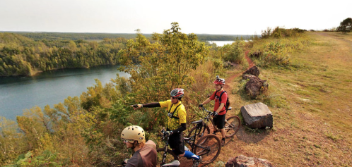 Three mountain bikers pause at a scenic overlook, admiring a river surrounded by lush greenery.