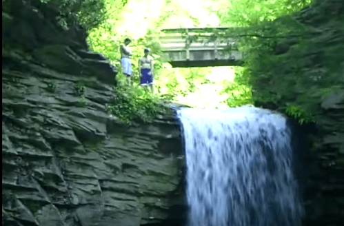 Two people stand on a rocky ledge near a waterfall, with a bridge visible in the background surrounded by greenery.