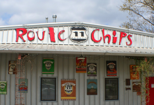 Sign for "Route 11 Potato Chips" on a metal building, with colorful posters and a cloudy sky in the background.
