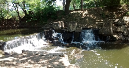 A serene waterfall cascades over stone steps into a calm river, surrounded by lush greenery and shaded trees.