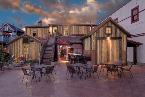 A rustic wooden building with outdoor seating, surrounded by trees, under a dramatic sky at dusk.