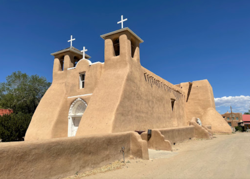Adobe-style church with two towers and crosses, set against a clear blue sky. Dusty road in the foreground.