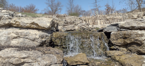 A small waterfall cascades over rocky terrain under a clear blue sky, surrounded by sparse vegetation.