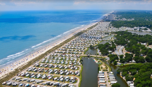 Aerial view of a beachside campground with rows of RVs, a winding river, and a coastline in the background.