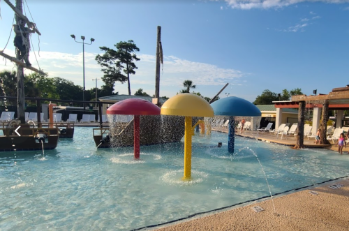 Colorful water play structures with fountains in a sunny pool area, surrounded by lounge chairs and trees.