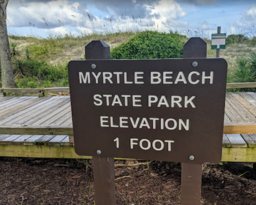 Sign at Myrtle Beach State Park indicating an elevation of 1 foot, with a wooden boardwalk in the background.