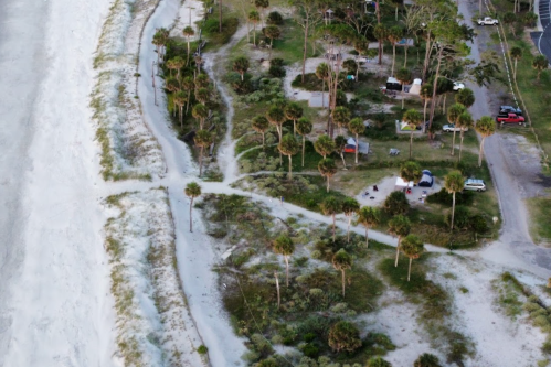 Aerial view of a beachside area with palm trees, pathways, and parked cars near a sandy shore.