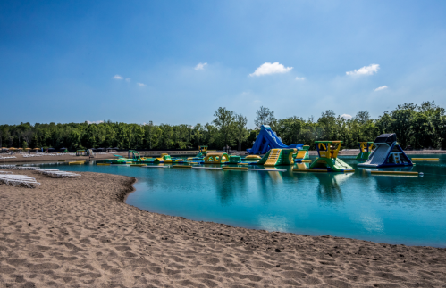 A sandy beach with a clear blue lake featuring inflatable water attractions and green trees in the background.