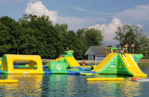 Colorful inflatable water park with slides and obstacles, surrounded by trees and a blue sky. People are enjoying the activities.