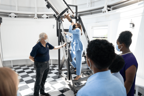 A group observes as a woman in a blue suit looks through a telescope, guided by a man in a black shirt.